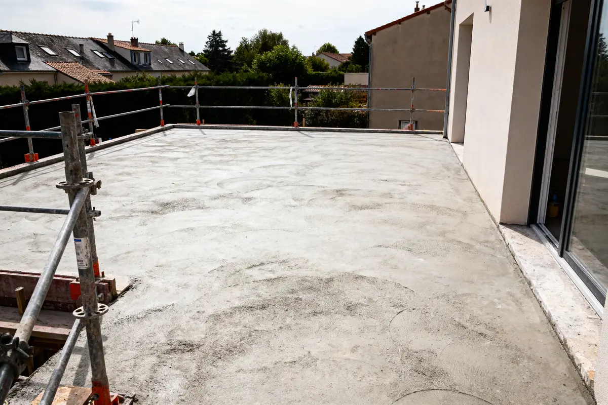 Terrasse en construction avec dalle béton, échafaudage, maisons voisines et ciel nuageux partiellement visible.