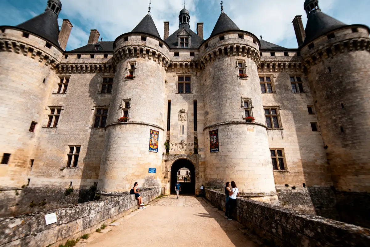 Château médiéval avec tours rondes, visiteurs assis et debout, ciel bleu.