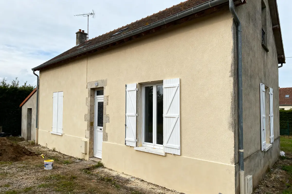 Petite maison beige avec volets blancs et antenne sur le toit, espace extérieur non aménagé.